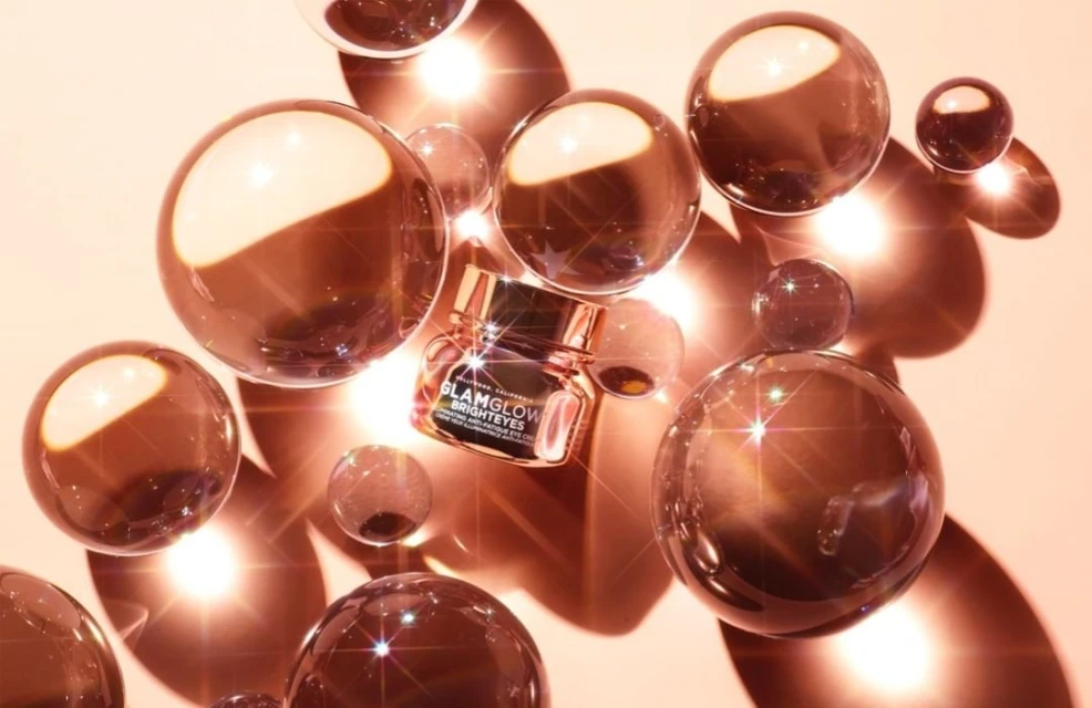 Glamglow Brighteyes eye cream jar with rose gold lid surrounded by transparent spheres on a peach background in studio lighting.