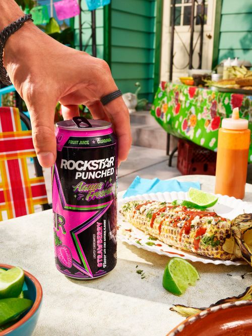 Person holding a can of Rockstar Punched Agua Fresca Strawberry drink outdoors with elotes, limes, and colorful decorations.