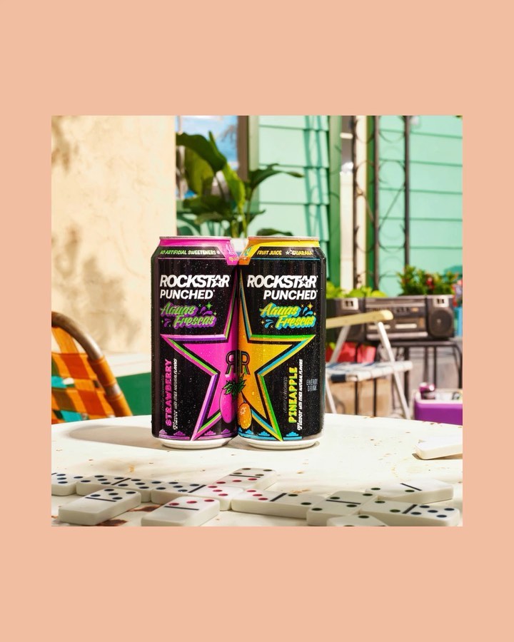 Two Rockstar Punched energy drink cans with strawberry and pineapple flavors on a tile table with dominoes, in an outdoor setting.
