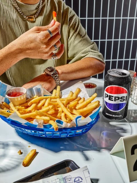 Close-up of a person eating french fries at a table with a can of Pepsi, featuring the diner setting and casual food photography.