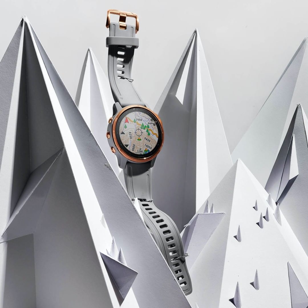 Smartwatch with grey band and rose-gold hardware is elevated against a white background of paper-folded origami mountains.