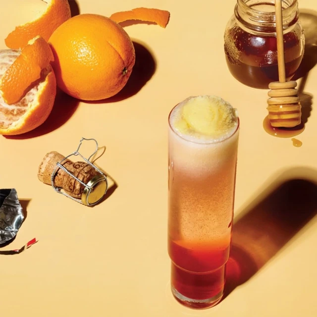 Tall glass filled with layered colorful cocktail, topped with yellow sorbet next to oranges and a honey dipper on a yellow surface.