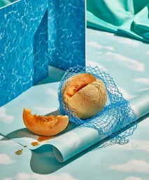 Cut cantaloupe melon wrapped in blue mesh net with a slice resting on a cloud-patterned surface beside a pool water backdrop.