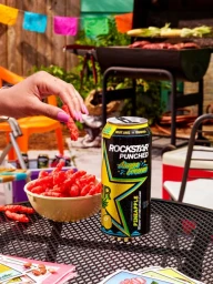 Rockstar Punched can and bowl of red snacks on a patio table with a hand holding one snack and a grill in the background.