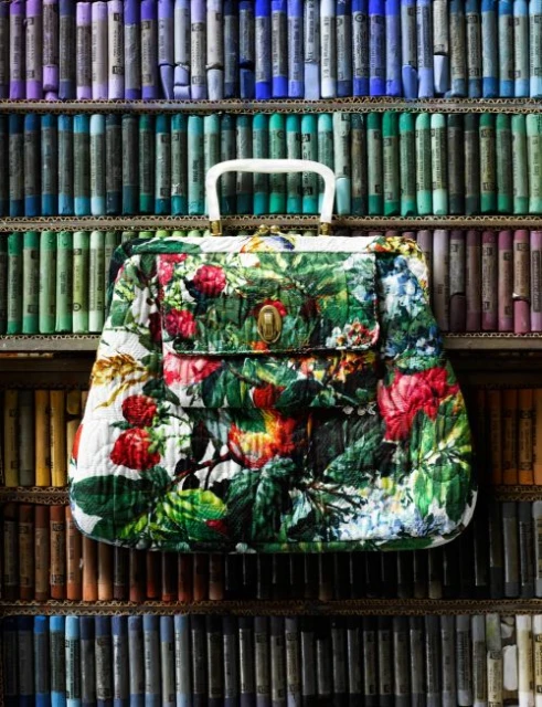 Quilted handbag with a floral pattern positioned in front of shelves lined with stacked, multi-colored pastel chalks.