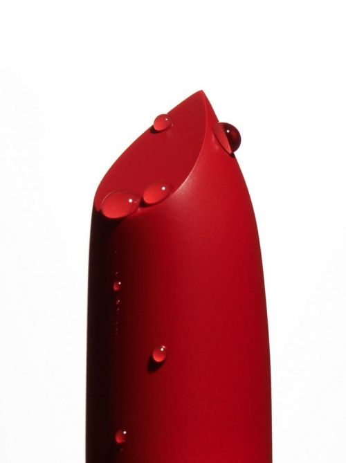 Macro studio shot of a red lipstick tube, adorned with water droplets against a clean white backdrop, showcasing cosmetic texture and color.