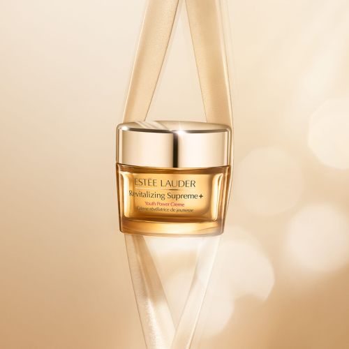 Close-up studio shot of Estee Lauder Revitalizing Supreme+ Youth Power Creme jar with gold lid and neutral ribbon accent.