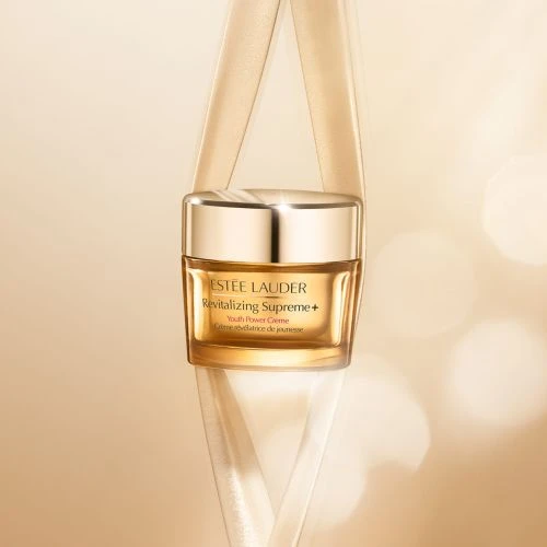 Close-up studio shot of Estee Lauder Revitalizing Supreme+ Youth Power Creme jar with gold lid and neutral ribbon accent.