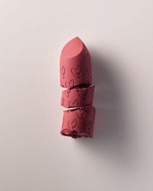 Close-up of broken pink lipstick with embossed heart pattern on a soft pink backdrop, showcasing texture and product detail.