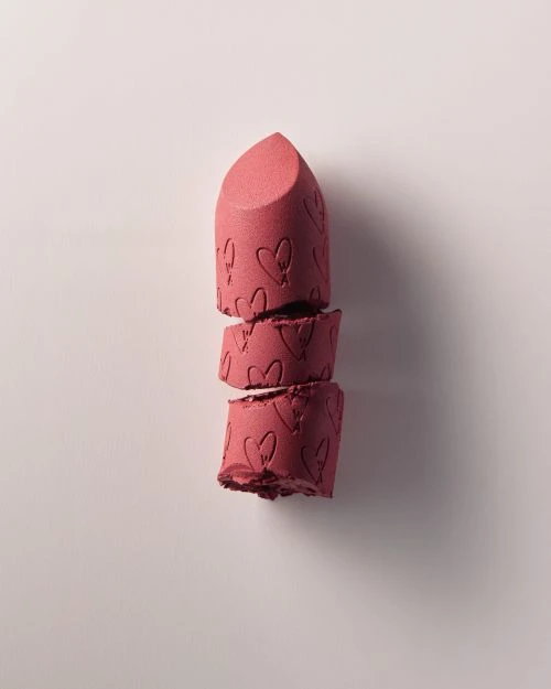 Close-up of broken pink lipstick with embossed heart pattern on a soft pink backdrop, showcasing texture and product detail.