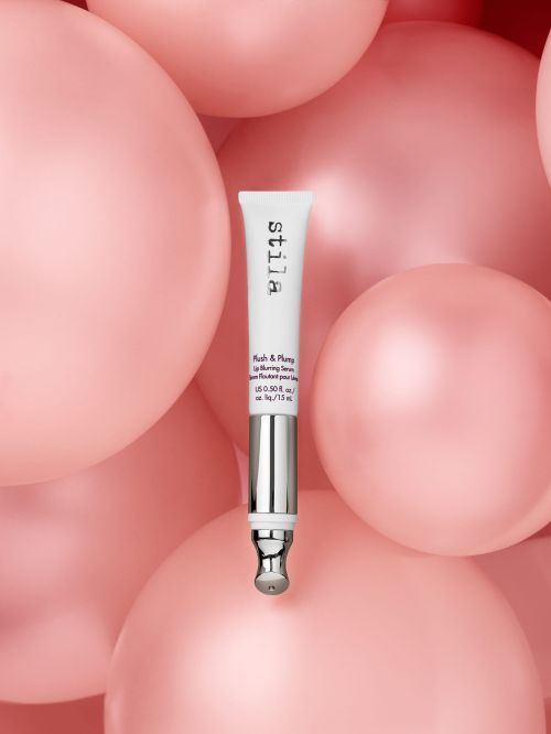 Close-up of a Stila lip serum tube in front of pink balloons, showcasing beauty product and cosmetic packaging.