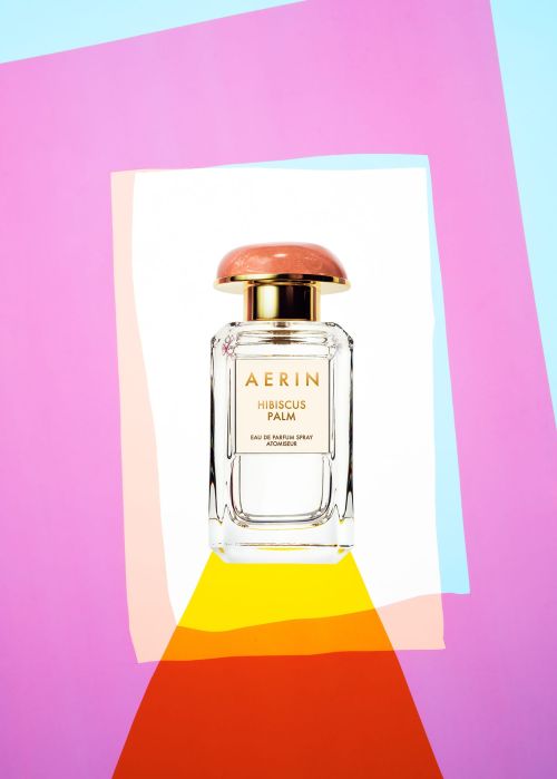 AERIN Hibiscus Palm perfume bottle sits centered on a white surface with colorful geometric shapes in the background.