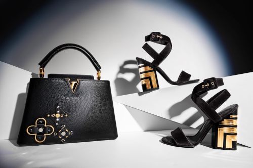 Fashion shot of a black leather handbag and a pair of black high-heeled sandals with gold geometric block heels on a white surface.