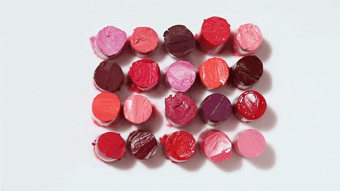 An overhead grid of twenty cut lipstick bullets in various shades of red, pink, and plum arranged on a white surface.