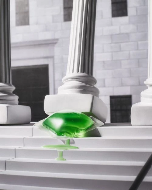 Green laundry detergent pod sits on a white set of steps with white columns in the background, in a product photography style.