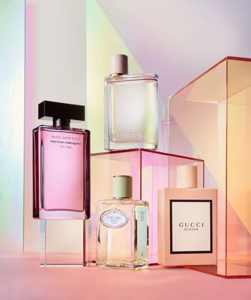 Four perfume bottles by Narciso Rodriguez, Burberry, Prada, and Gucci arranged on clear blocks against a gradient pink background.