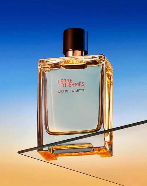 Square-shaped Terre d'Hermès Eau de Toilette perfume bottle on a transparent surface with a gradient blue to orange background.