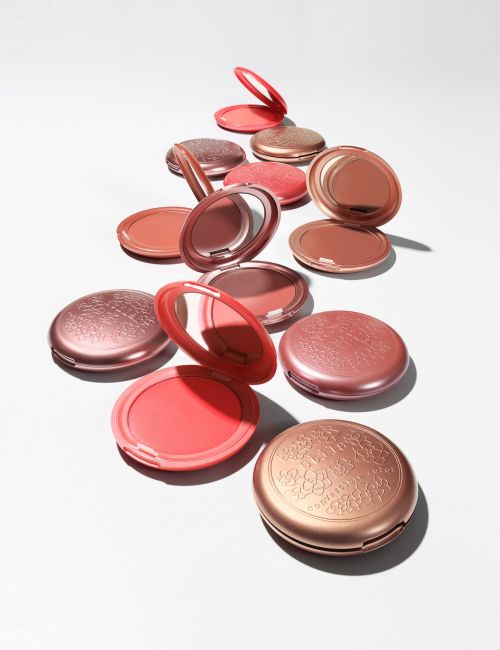 Arrangement of pink and coral-toned makeup compacts with decorative lids and mirrors, displayed on a white surface in studio lighting.