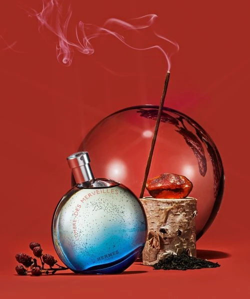 Round perfume bottle on wooden block with amber stone, lit incense, red background. Aromatherapy and fragrance product photography.