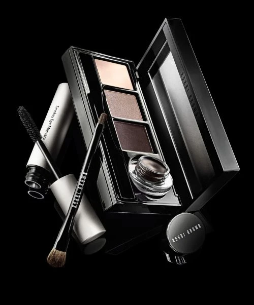Bobbi Brown makeup products including an eye shadow palette, mascara, eye liner pot, and makeup brush against a dark background.