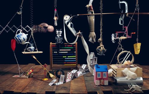 Still life arrangement with miniature house, playing cards, teapot, abacus, and suspended objects against a dark background.
