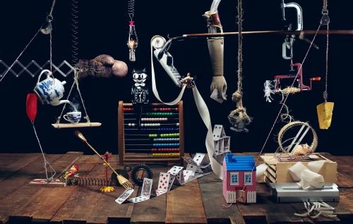Still life arrangement with miniature house, playing cards, teapot, abacus, and suspended objects against a dark background.
