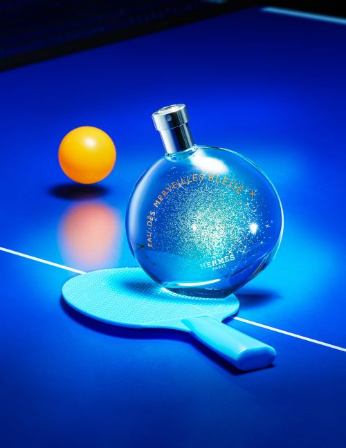 Perfume bottle by Hermès sits on a ping pong paddle and table beside a ping pong ball, all lit with a blue light.