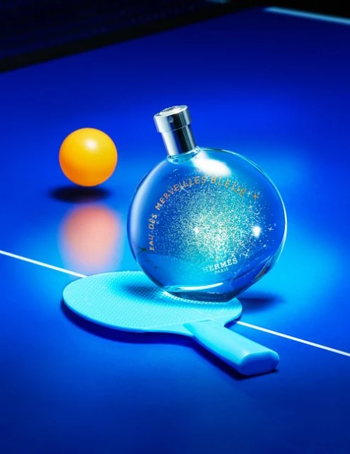 Perfume bottle by Hermès sits on a ping pong paddle and table beside a ping pong ball, all lit with a blue light.