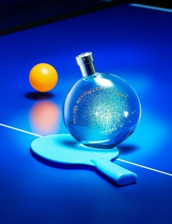 Perfume bottle by Hermès sits on a ping pong paddle and table beside a ping pong ball, all lit with a blue light.