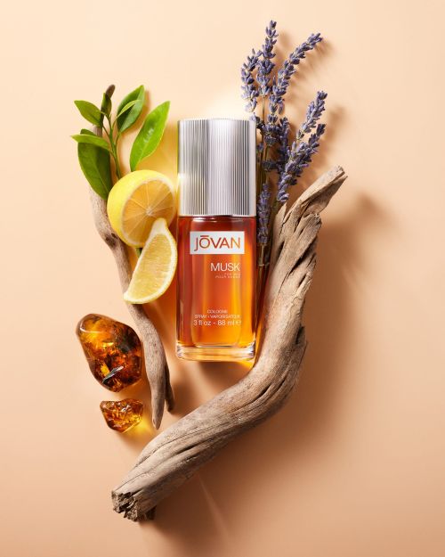 Still life of Jovan Musk cologne bottle surrounded by botanical elements: lemons, lavender, amber, and weathered driftwood on peach backdrop.