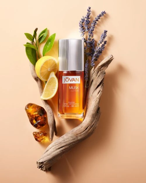 Still life of Jovan Musk cologne bottle surrounded by botanical elements: lemons, lavender, amber, and weathered driftwood on peach backdrop.