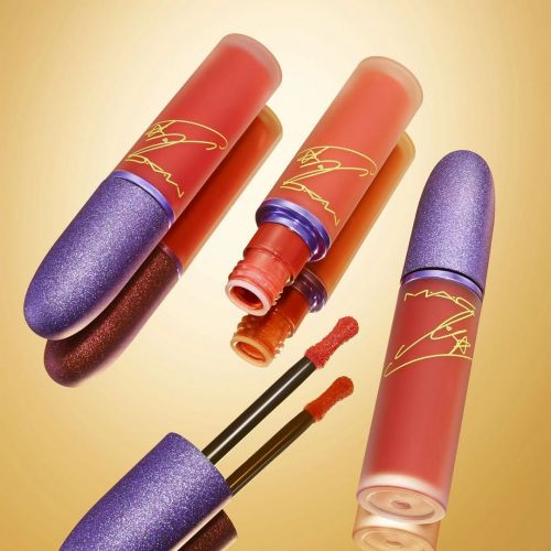 Three MAC lipsticks with purple caps are angled on a reflective surface in a beauty-style product shot with a soft, warm-toned background.