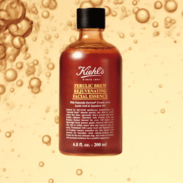 Kiehl's Ferulic Brew Rejuvenating Facial Essence bottle in amber liquid against a blurred golden background with oil droplet patterns.