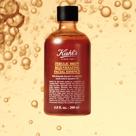 Kiehl's Ferulic Brew Rejuvenating Facial Essence bottle in amber liquid against a blurred golden background with oil droplet patterns.