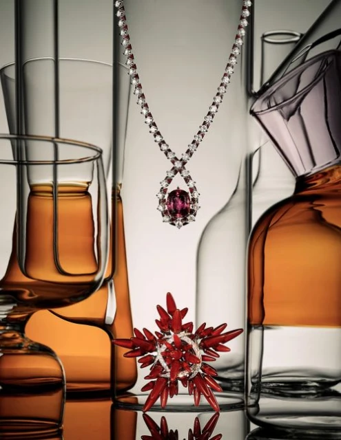 A necklace with a ruby pendant is the focal point, surrounded by clear and amber-tinted glassware creating a warm and reflective aesthetic.