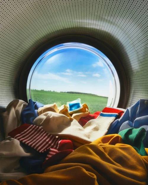 Clothes and socks tumbling inside an open washing machine drum, against a background of a bright blue sky and green field.