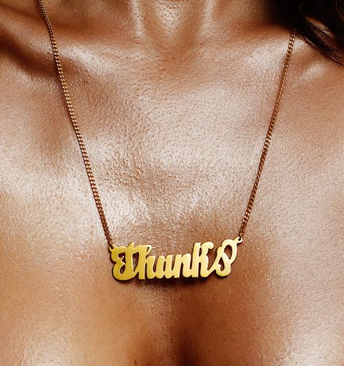 Close-up shot of an adult female wearing a gold chain necklace with the word 'Thanks' hanging at the center. Fashion shot.