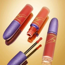 Three lipstick tubes with purple sparkly caps and peach red bodies arranged diagonally on a reflective golden surface.