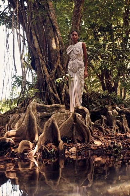 Woman in a draped gown poses in a lush forest setting with exposed tree roots and still water reflecting the verdant foliage.