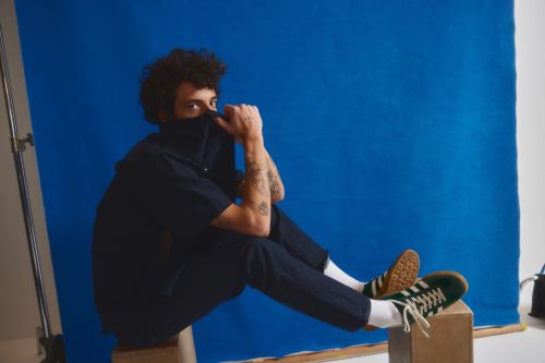Portrait of a curly-haired person in a navy outfit sitting on a wooden box, holding their collar and wearing green and brown sneakers.