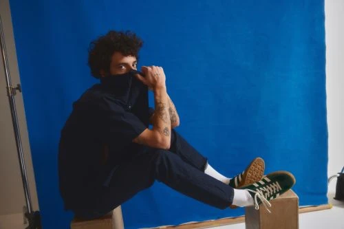 Portrait of a curly-haired person in a navy outfit sitting on a wooden box, holding their collar and wearing green and brown sneakers.