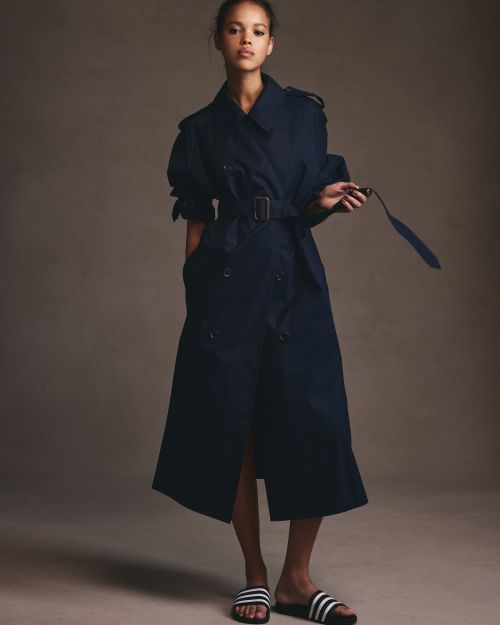 A woman poses in a studio, wearing a navy trench coat and athletic slides, against a neutral backdrop, showcasing a fashion-forward style.