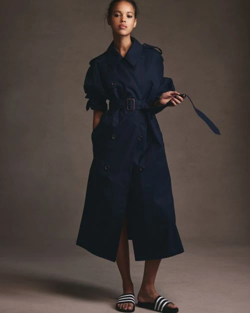 A woman poses in a studio, wearing a navy trench coat and athletic slides, against a neutral backdrop, showcasing a fashion-forward style.