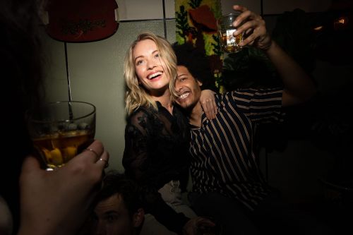 Blonde woman and man with dark curly hair at a bar, holding up glasses with alcohol in them, smiling; dimly lit and close-up shot.