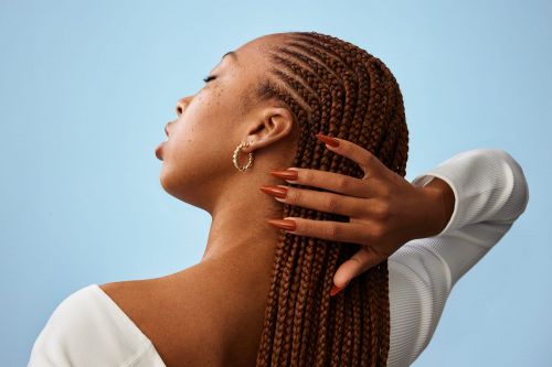 Close-up of a young Black woman with brown braided hair and long orange nails against a light blue backdrop in a studio shot.