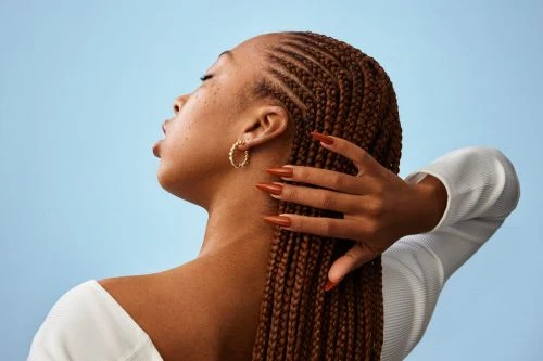 Close-up of a young Black woman with brown braided hair and long orange nails against a light blue backdrop in a studio shot.