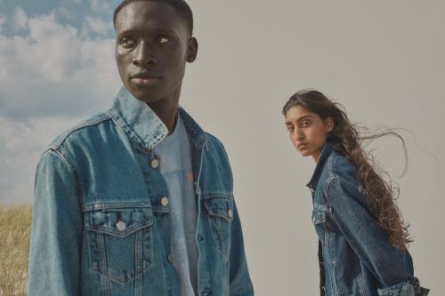 Two fashion models wearing denim jackets pose outdoors against a bright sky. A modern fashion advertisement for clothing.