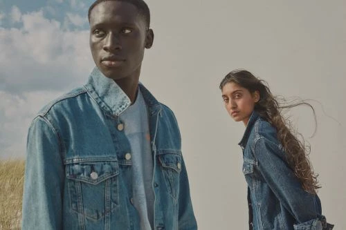 Two fashion models wearing denim jackets pose outdoors against a bright sky. A modern fashion advertisement for clothing.