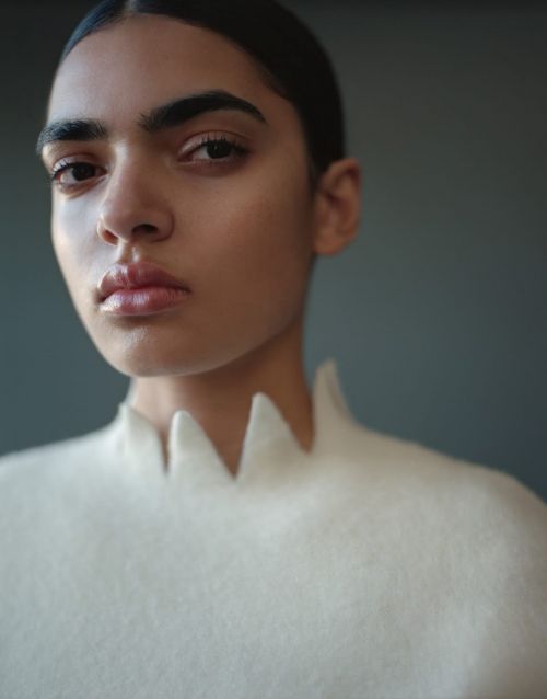 Close-up studio portrait of a young adult female with dark hair, wearing a textured white top against a neutral gray background.