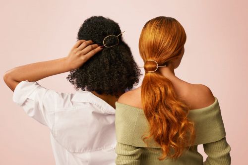 Two women with different hair colors stand close together showcasing gold hairpins in a studio with a soft pink background.